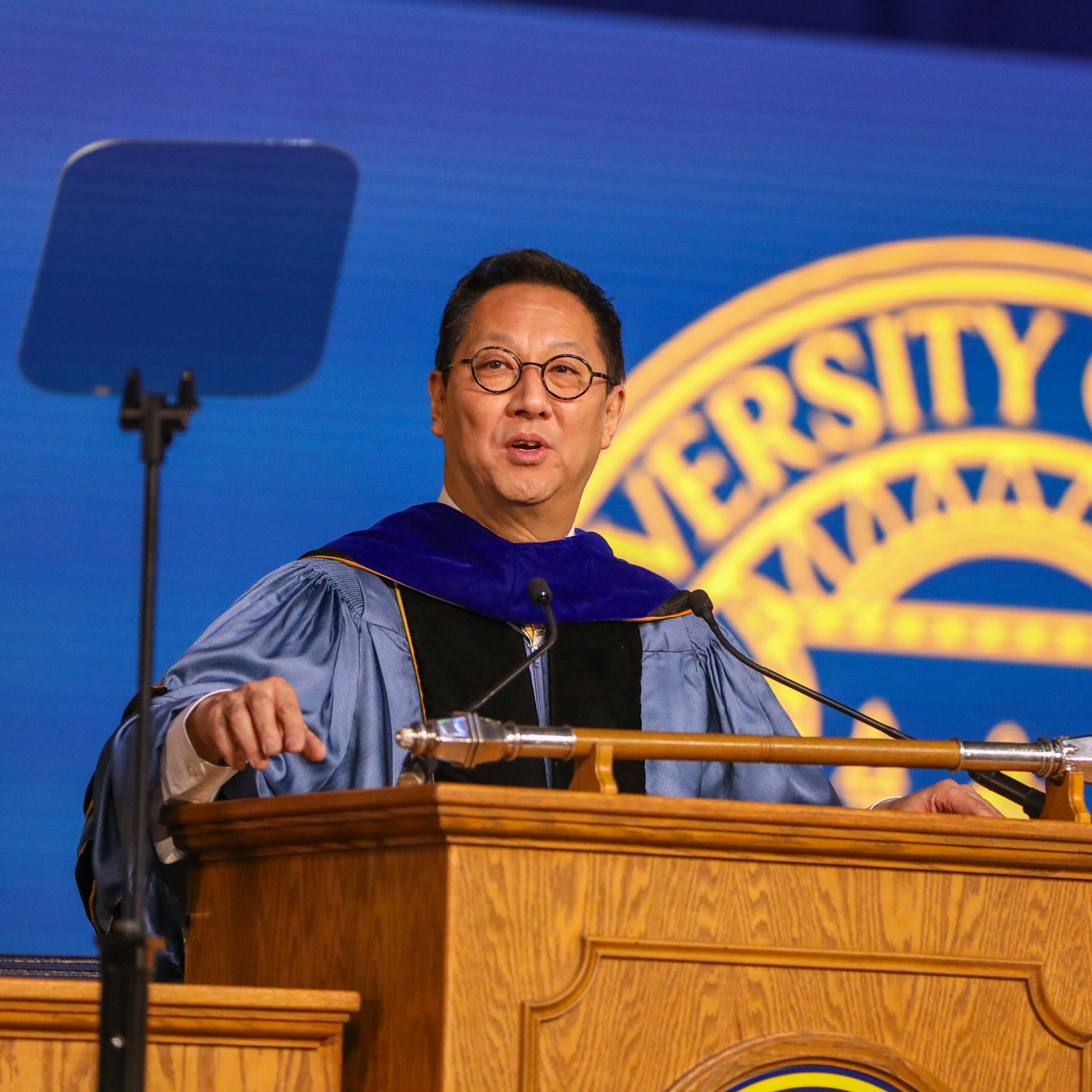 Santa Ono, in graduation robes, speaks at a brown podium with a U-M logo projected behind him.