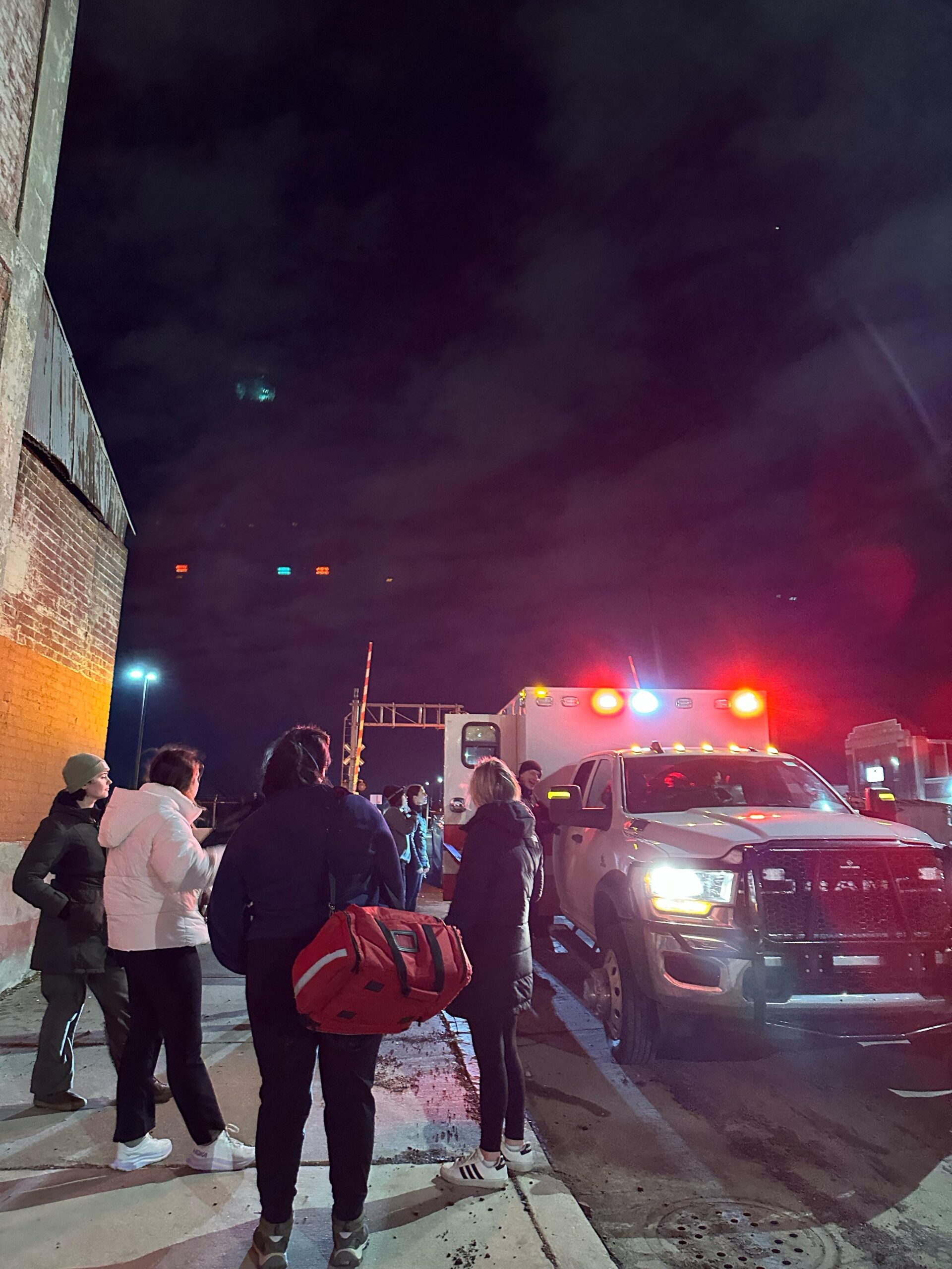 Wolverine Street Medicine volunteers and EMTs standing around an ambulance with its flashing lights on in a night time scene.