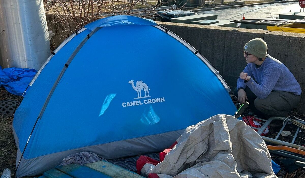A Wolverine Street Medicine volunteer kneels outside a blue tent positioned in a urban sidewalk area.