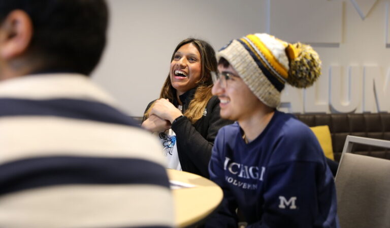 Two students, a male and female, laugh while looking forward. The male student is wearing a University of Michigan stocking hat.