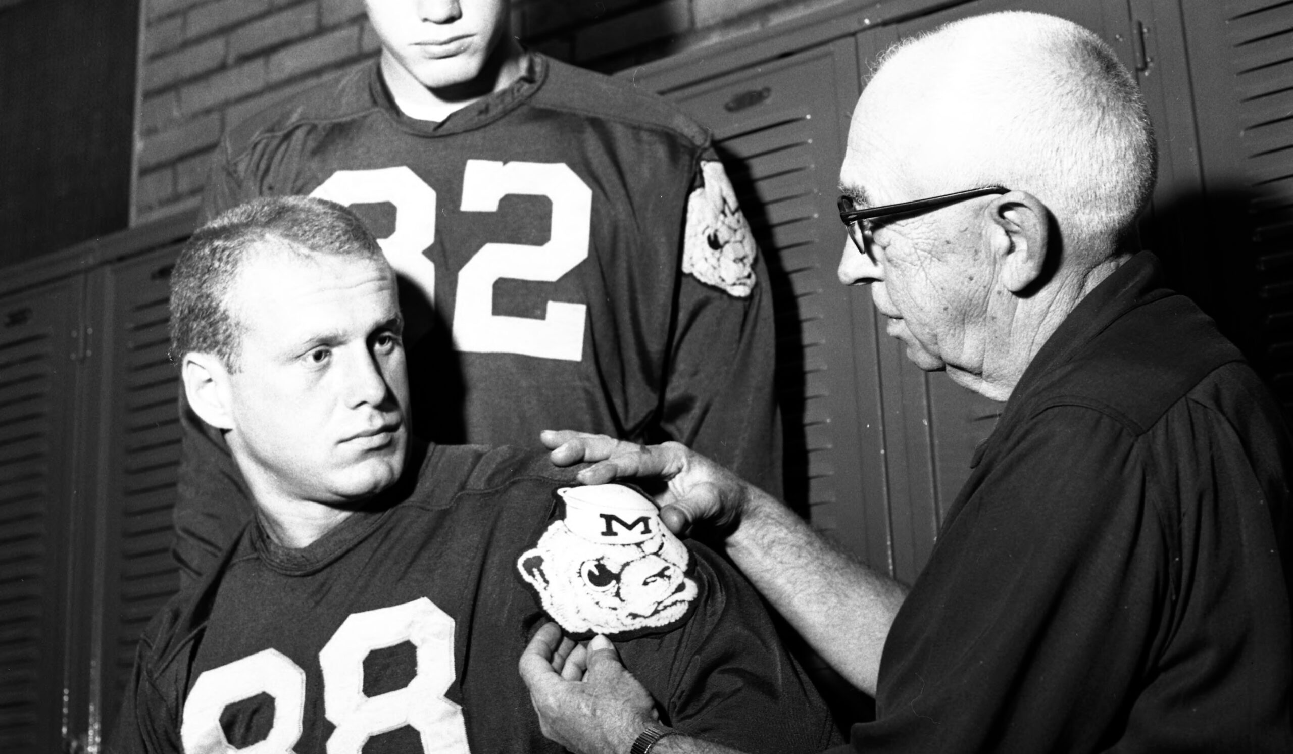 An older man in glasses places a wolverbear logo on the sleeve of a football player wearing number 88. The photo is in black and white.