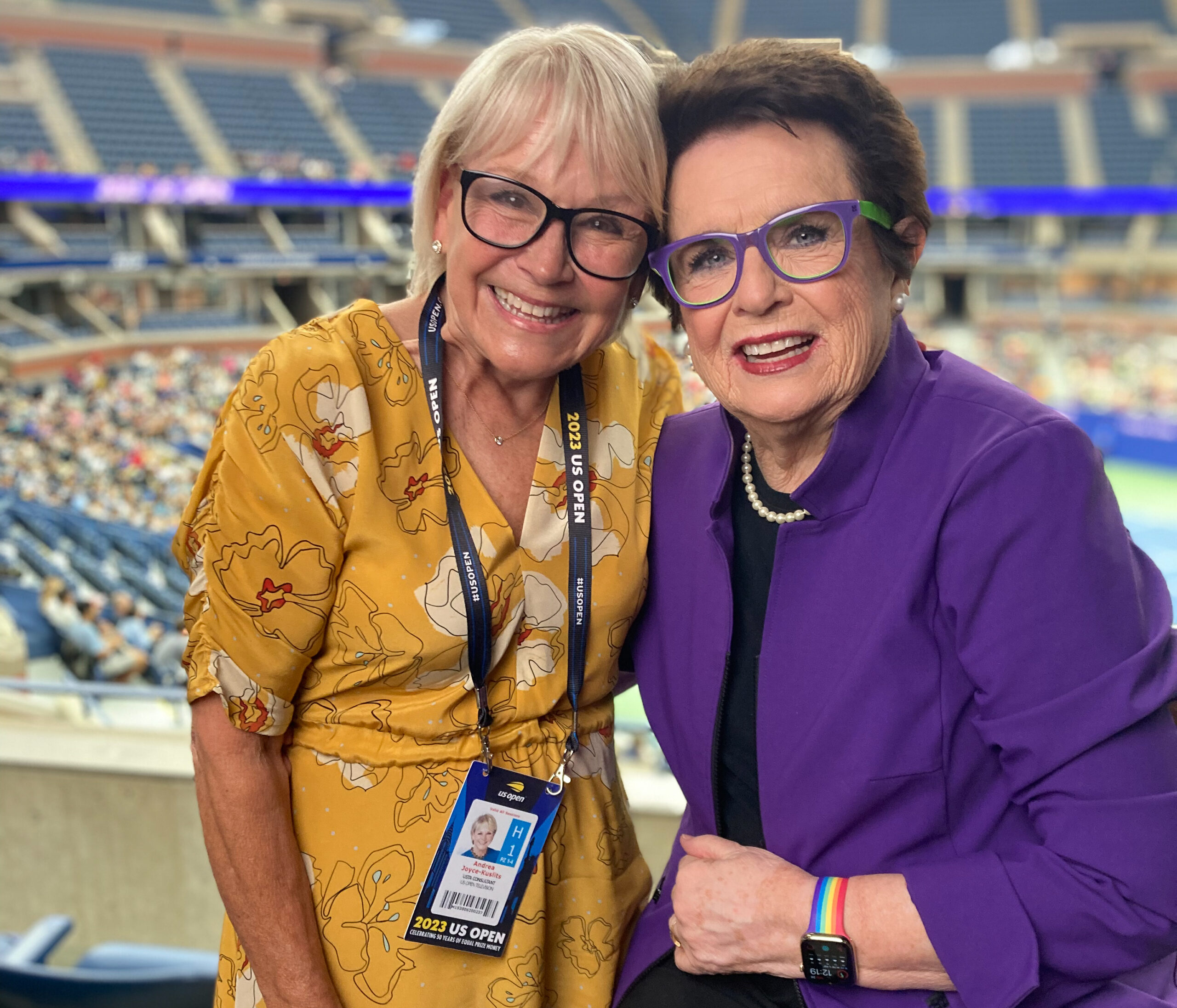 Joyce, left, in a yellow dress, poses with her arm around Billie Jean King, right, who is wearing a black top and purple dress jacket.