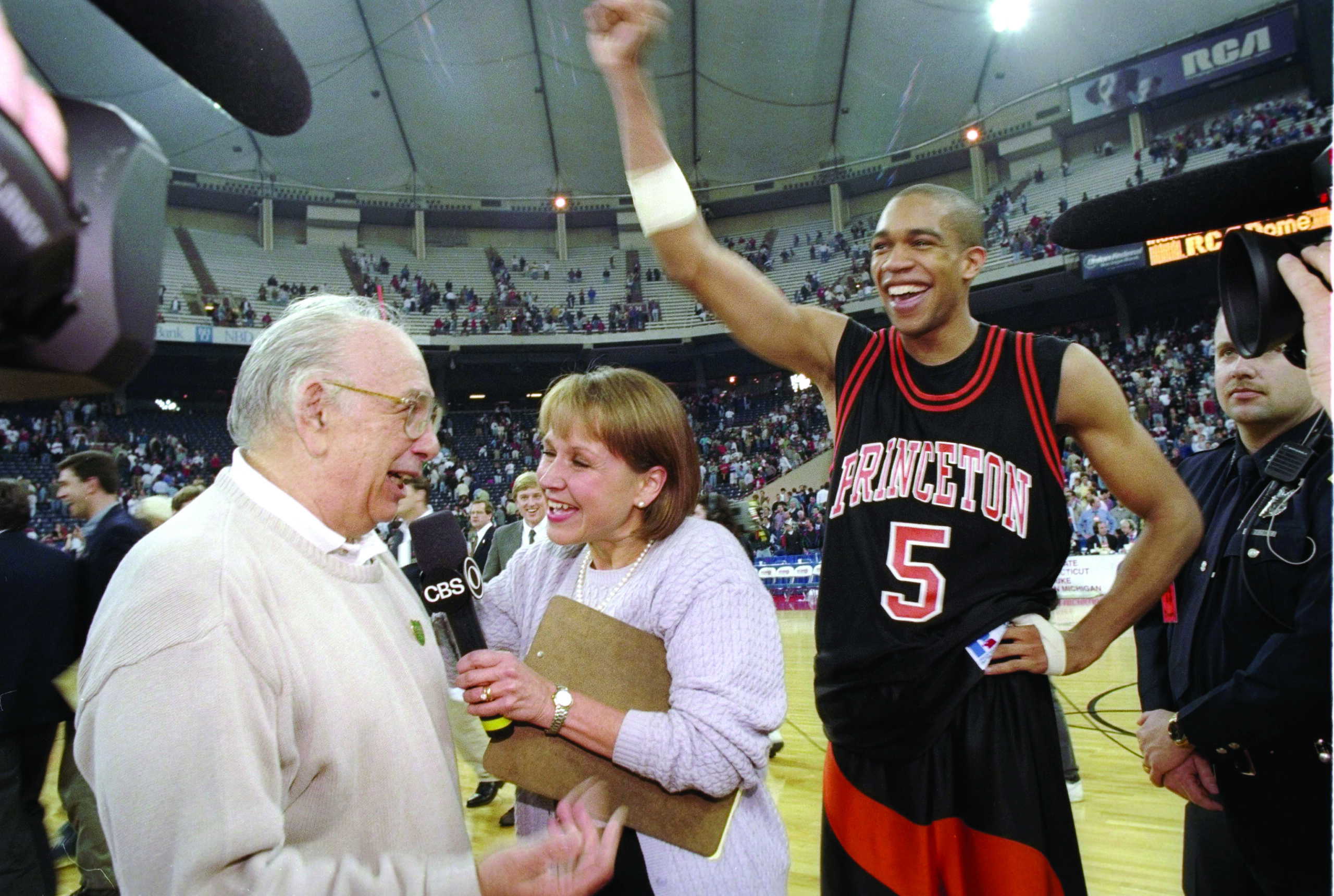 Pete Carril, left, in a tan sweater, speaks into a microphone held by Andrea Joyce, center. Right is Sydney Johnson in a Princeton basketball uniform. They are standing in a basketball arena.