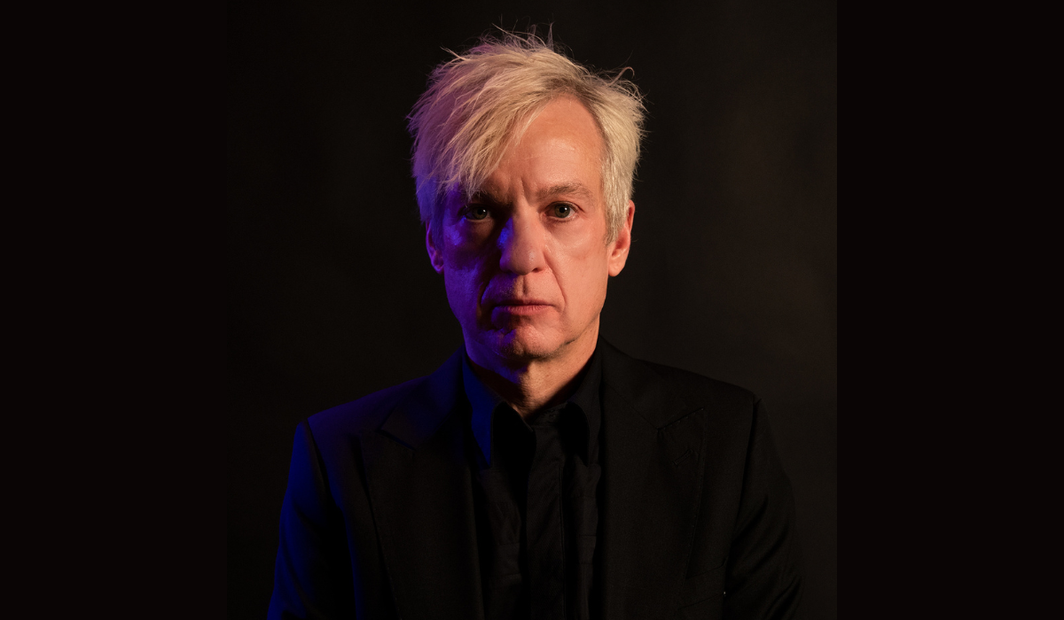 Chris Vrenna, with short blond hair, poses seriously for a headshot in a black shirt and dark room. There is a slight blue/purple glow on the left side of his face.