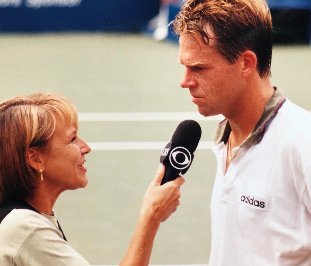 Joyce, left, holds a black CBS microphone up to Stefan Edberg, right, who is visibly sweating and wearing a white and black Adidas polo shirt. There is a tennis court in the background.