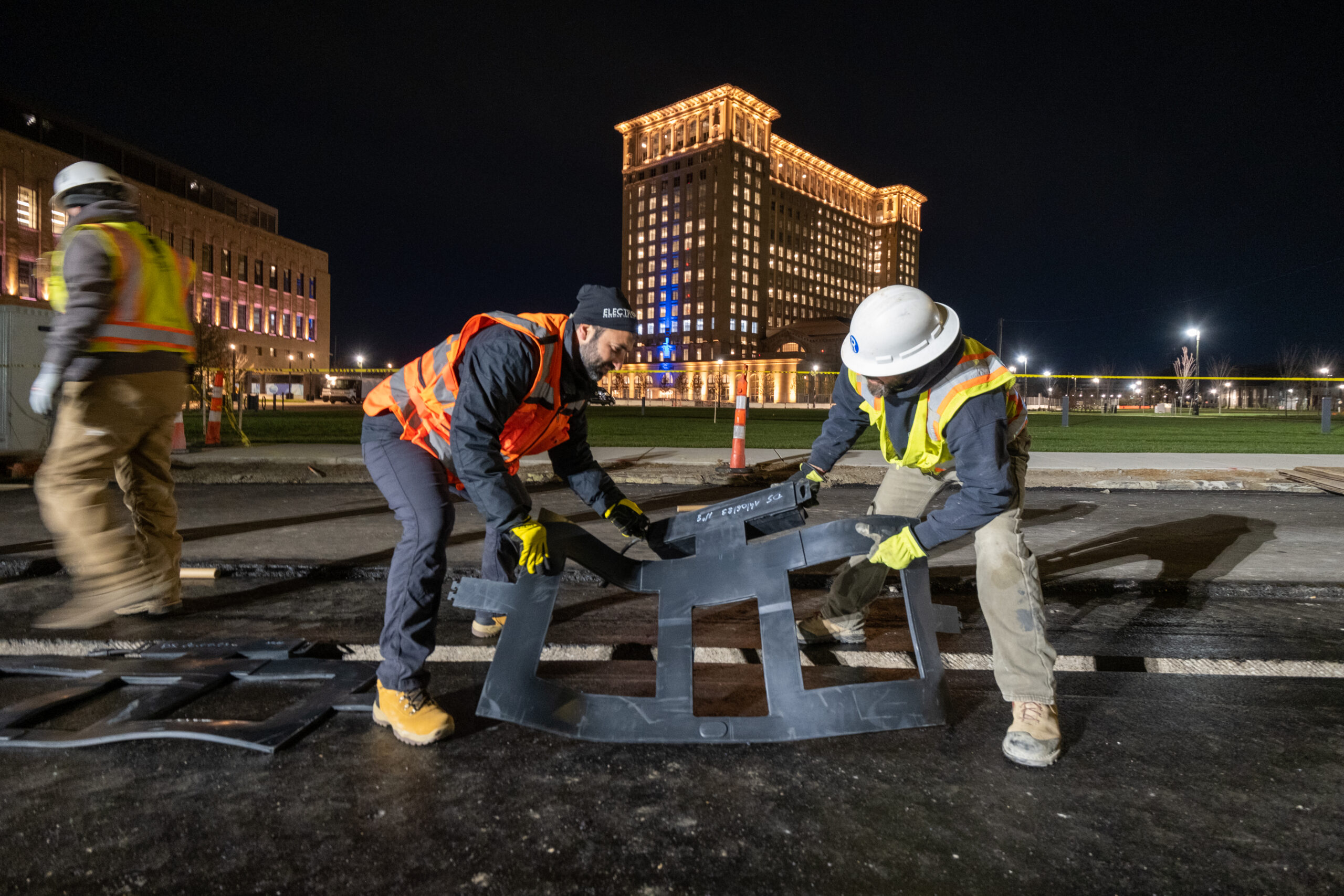 Two people in hard hats lay charging pieces during road construction. It's nighttime and Michigan Central Station is illuminated in the background.