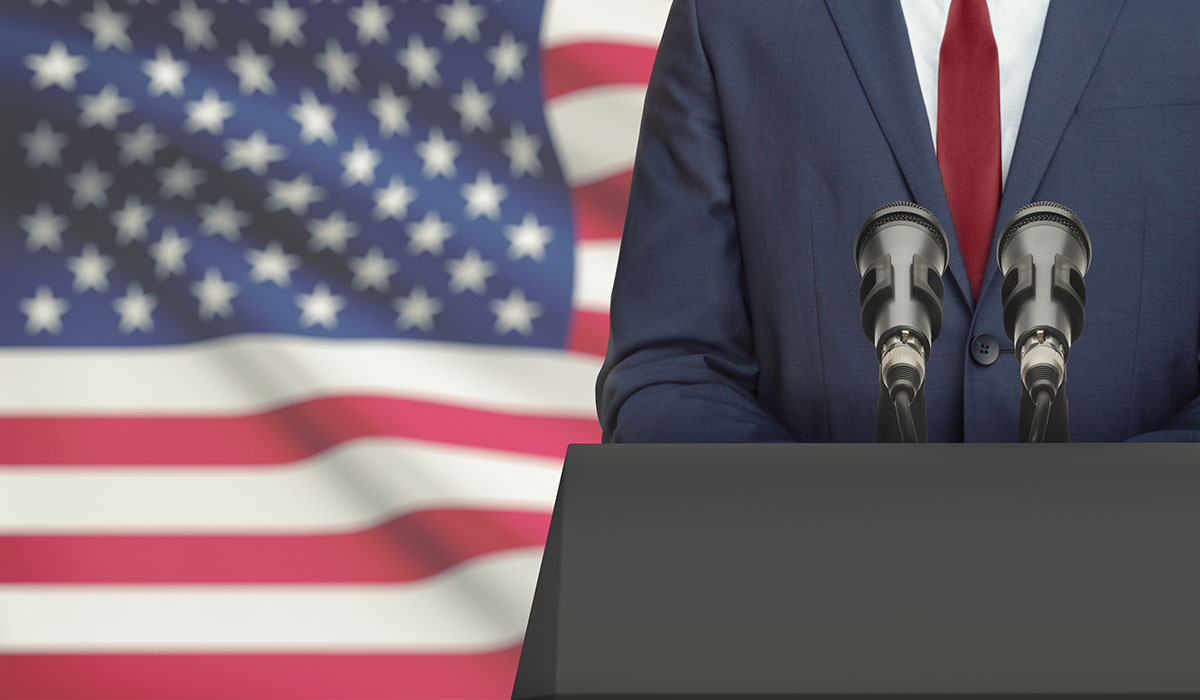 Businessman Or Politician Making Speech From Behind A Pulpit With National Flag On Background United States