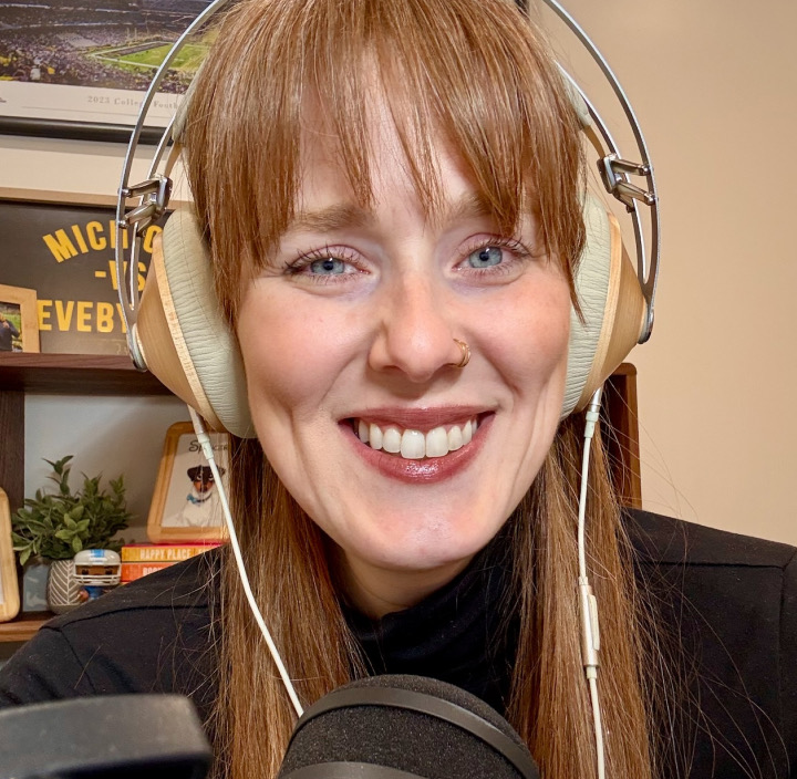 A close-up headshot of Kristy, who is smiling and wearing headphones with a mic in front of her.