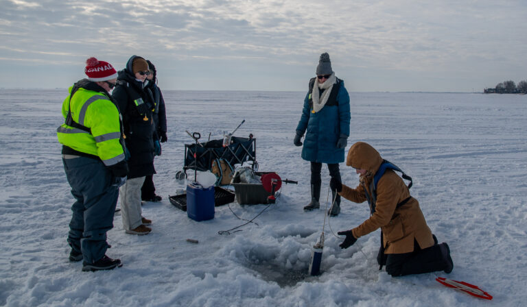 Researchers in winter clothing conduct winter water sampling in a hole in Lake St. Clair. There is snow on the ice.