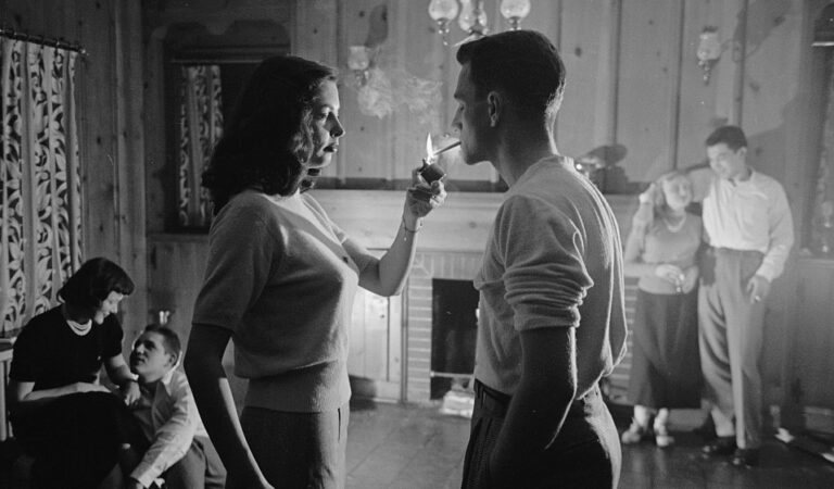 A black and white image from the 1940s of a woman (left) lighting a man's cigarette (right) with a fireplace in the background.
