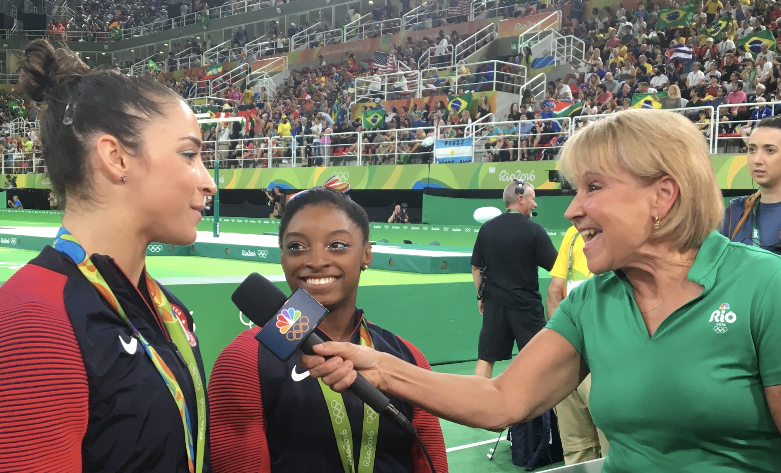 Joyce, far right, in a green Rio Olympics polo shirt, holds a microphone to Aly Raisman, far left, who is in a USA top and wears a medal around her neck. Simone Biles, in the same outfit, stands in the middle smiling at Raisman.