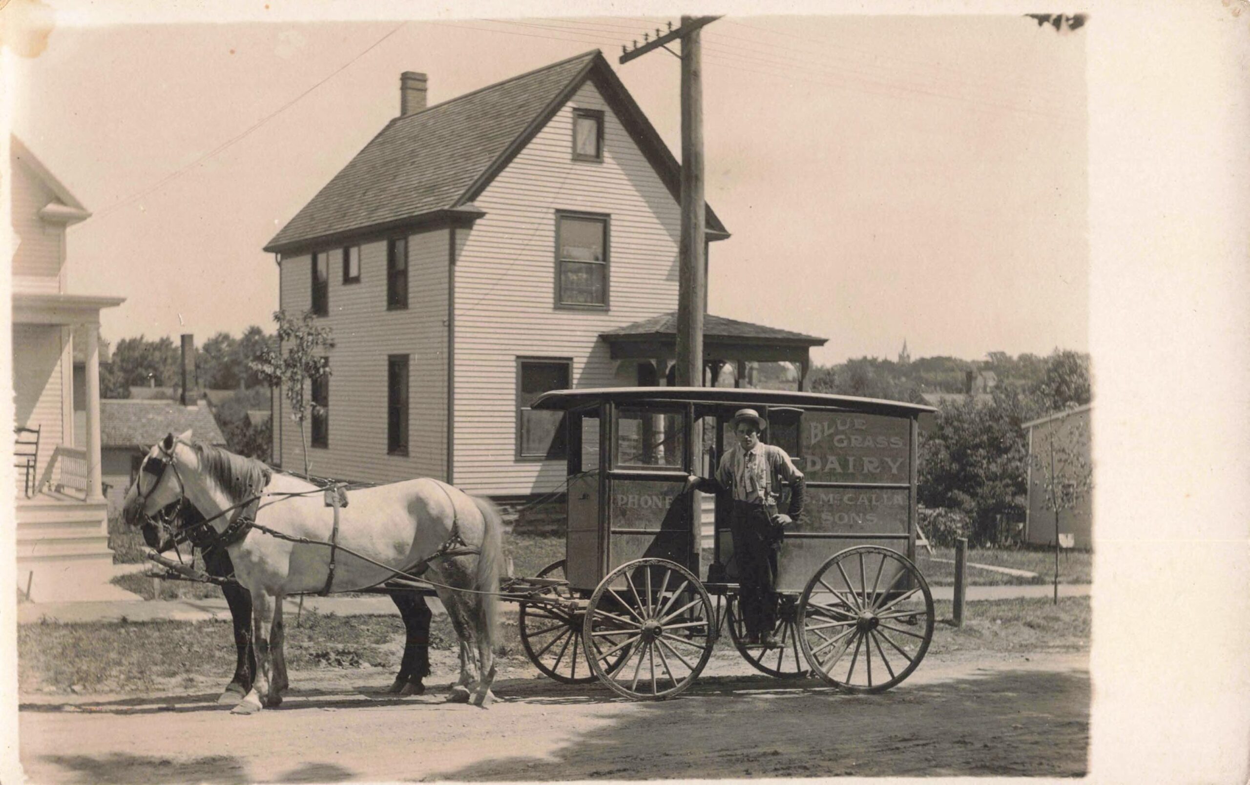 A sepia-toned photo of a horse drawn carriage delivering milk and a farm house behind them.