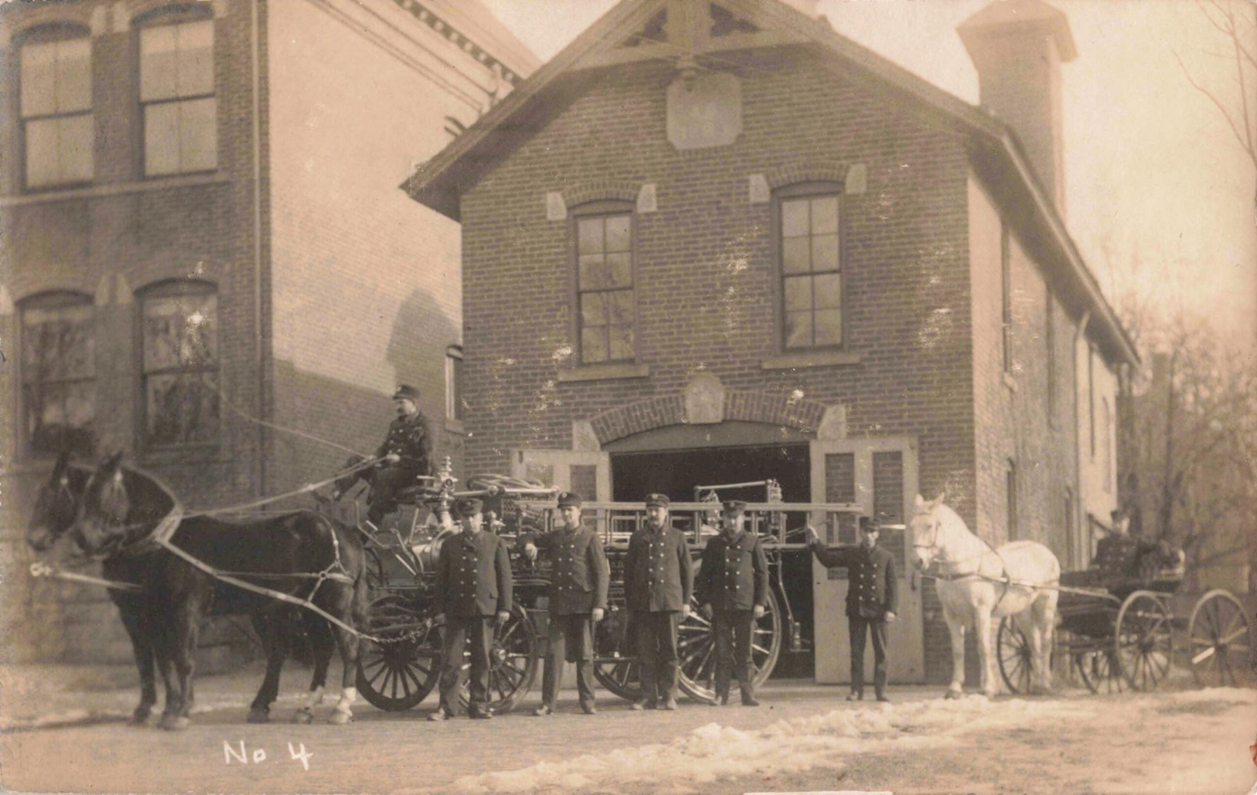 Five people in dark uniforms stand in front of two horse drawn carriages with a brick fire house in the background.
