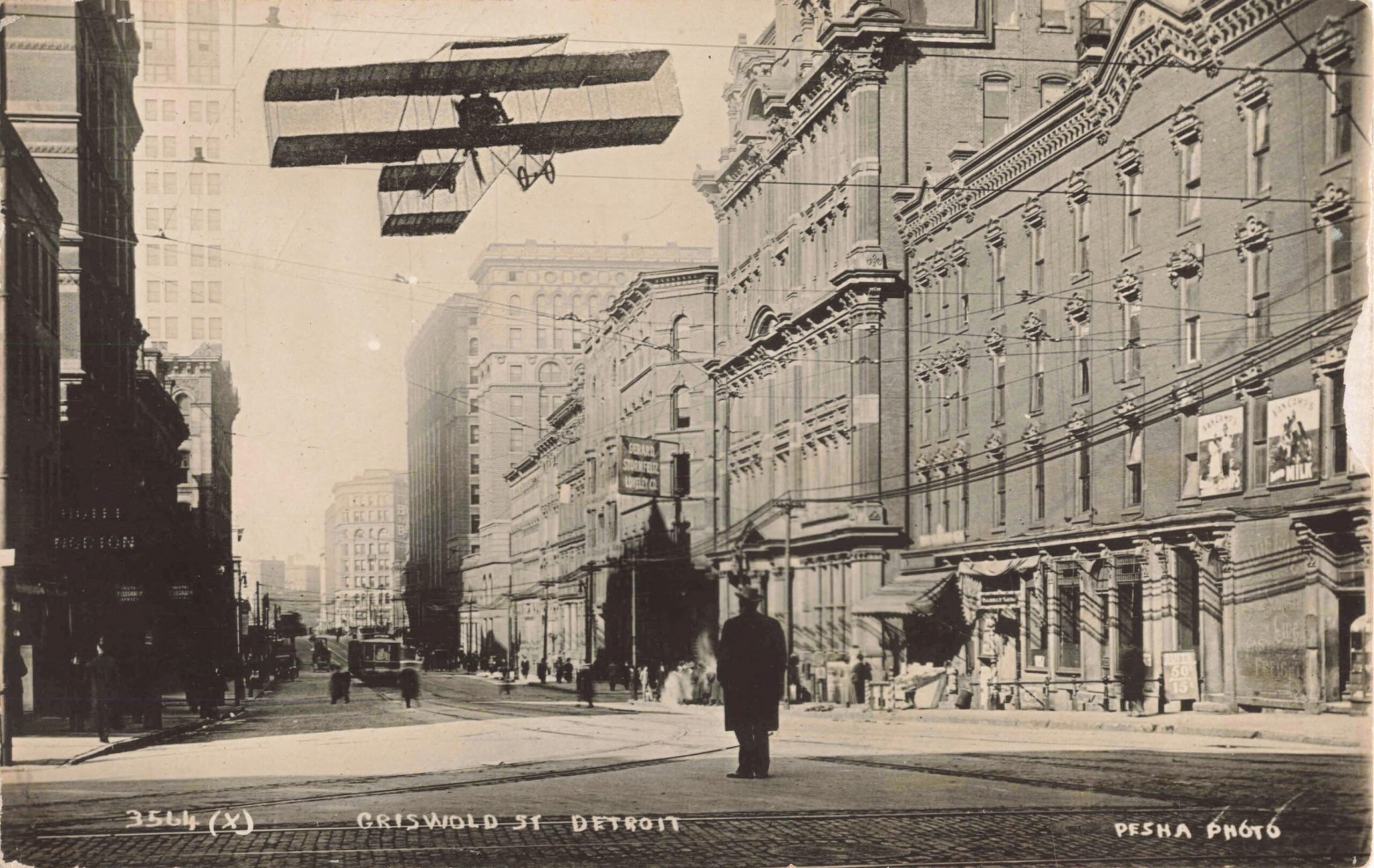 Black and grey postcard image depicting Griswold Street in Detroit in 1910. There is a low-flying airplane and a nondescript individual standing in the middle of the street.