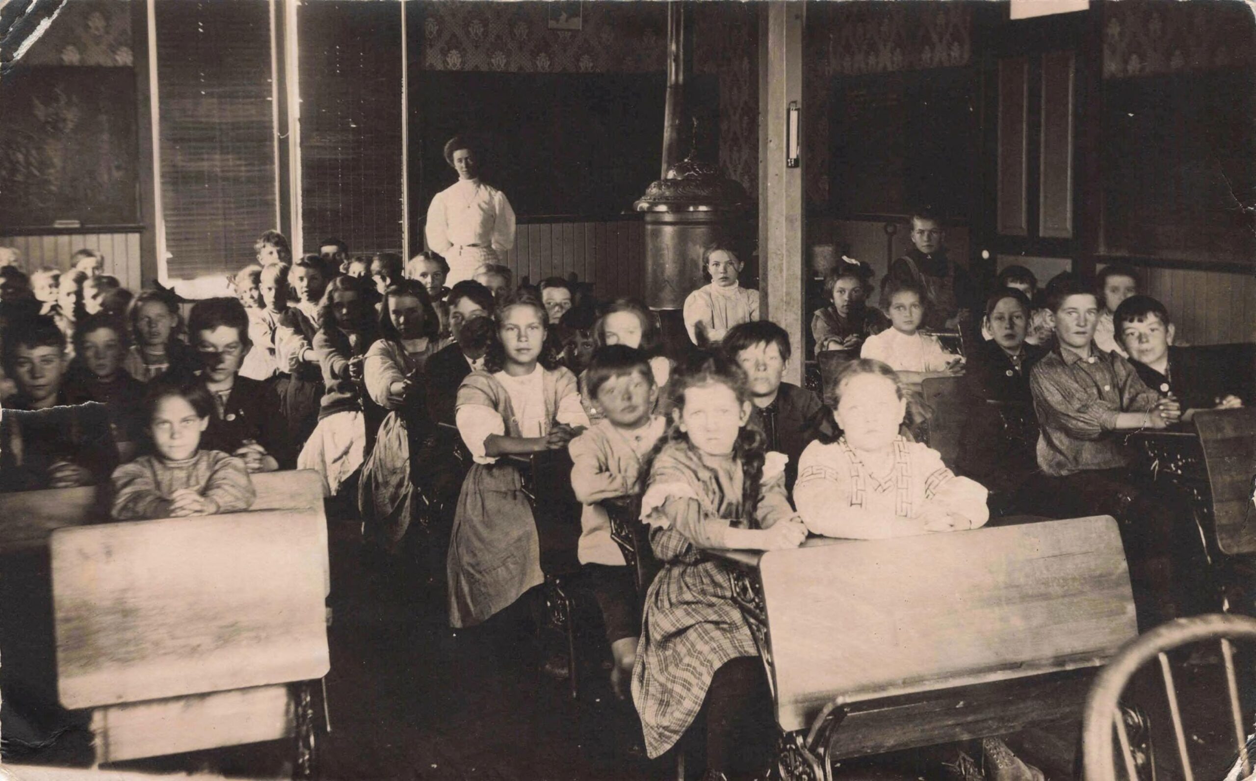 A sepia-toned image of an old schoolroom in 1908 with many children at close desks, many children two to a desk.