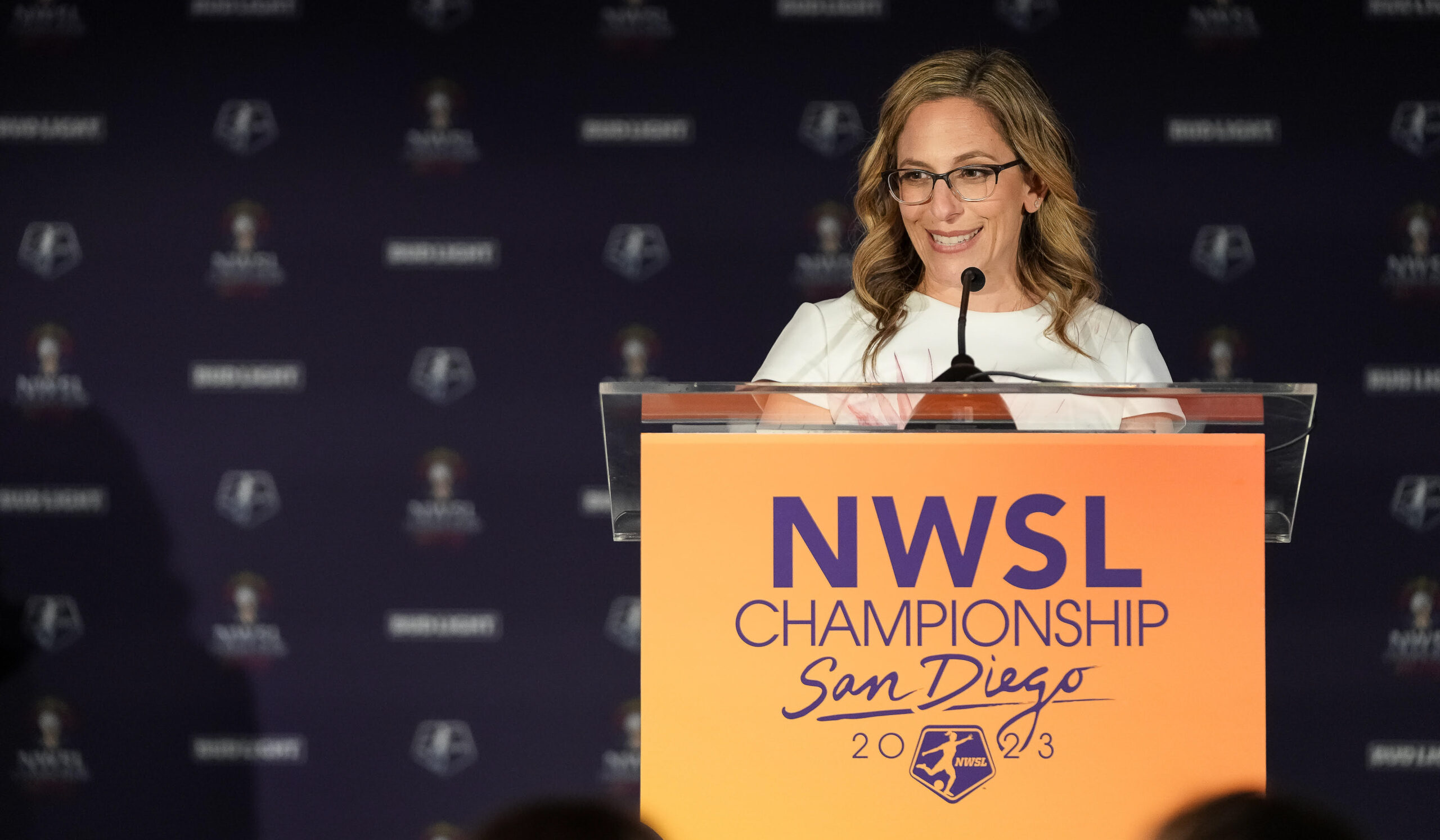 Jessica Berman, in a white top, stands in front of an orange podium with the words "NWSL Championship San Diego 2023."