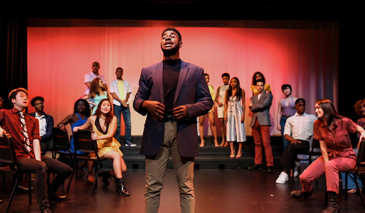 One student in a suit jacket and khaki pants sings center stage with a group of other students around him. The background is red.