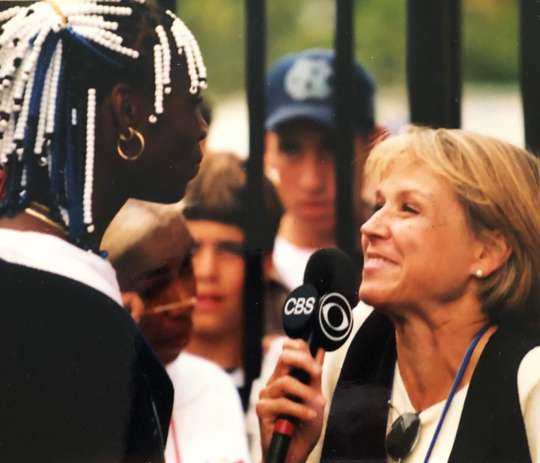 Venus Williams, left, with white and blue beads in her hair, looks at Joyce, left, who is holding a CBS microphone to her mouth. People watch them in the background behind a tall fence.