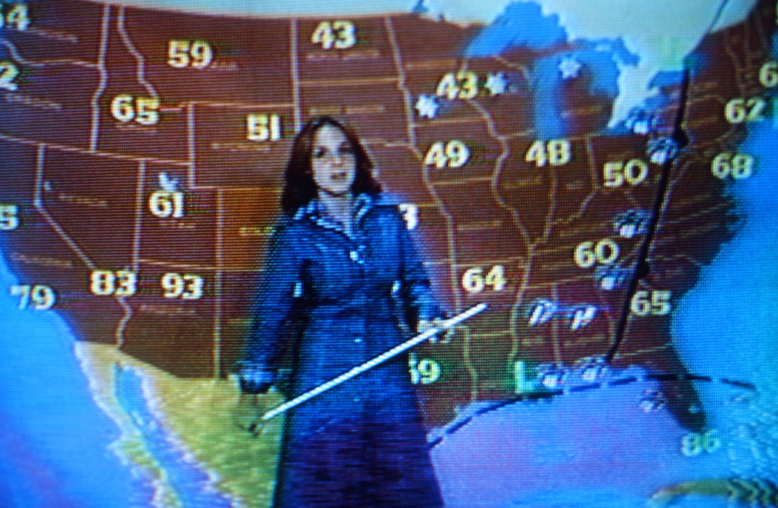 An early 1980s photo of Andrea Joyce in a jean dress with a pointer stick in front of a temperature map as a weather person.